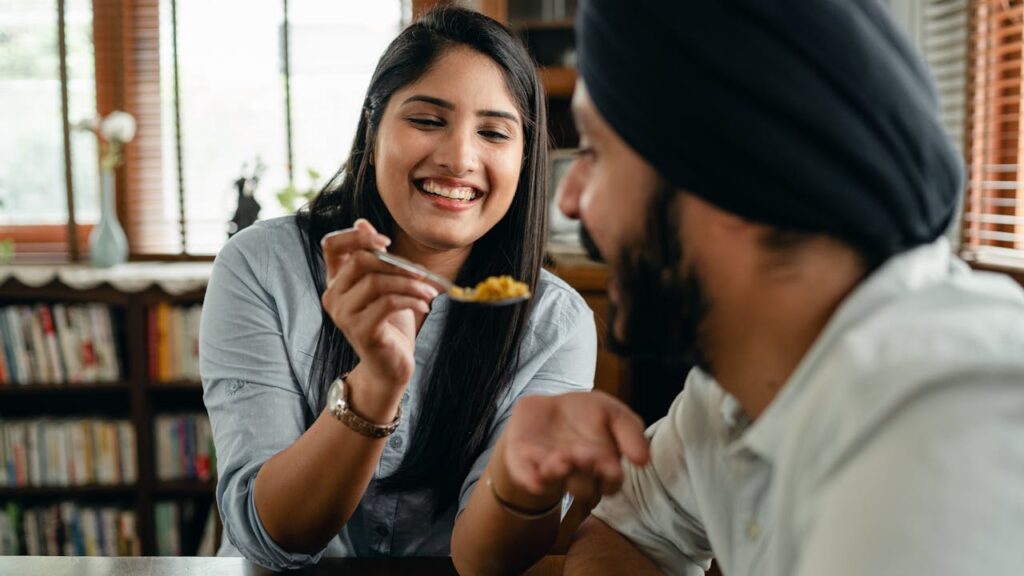 Happy young Indian woman smiling and feeding positive hungry boyfriend with delicious saffron rice